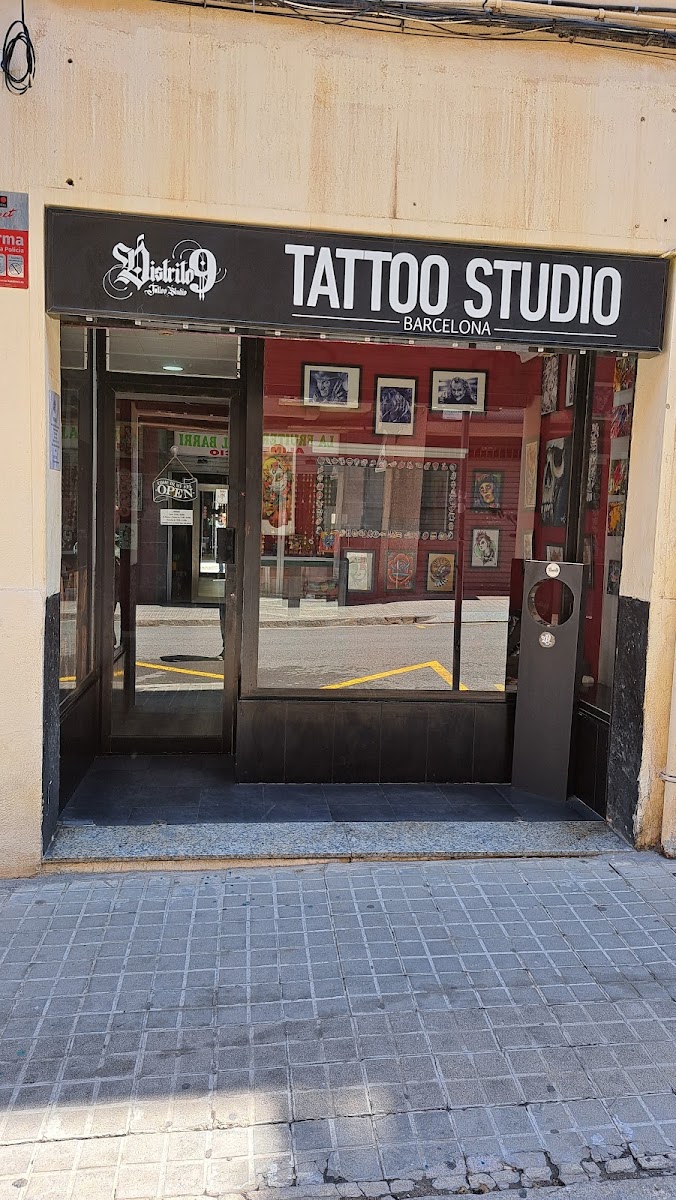 Fachada del estudio: rótulo negro con Distrito 9 en letra gótica y las palabras TATTOO STUDIO BARCELONA, sobre un escaparate oscuro con la pared roja del interior y láminas enmarcadas al otro lado del cristal.