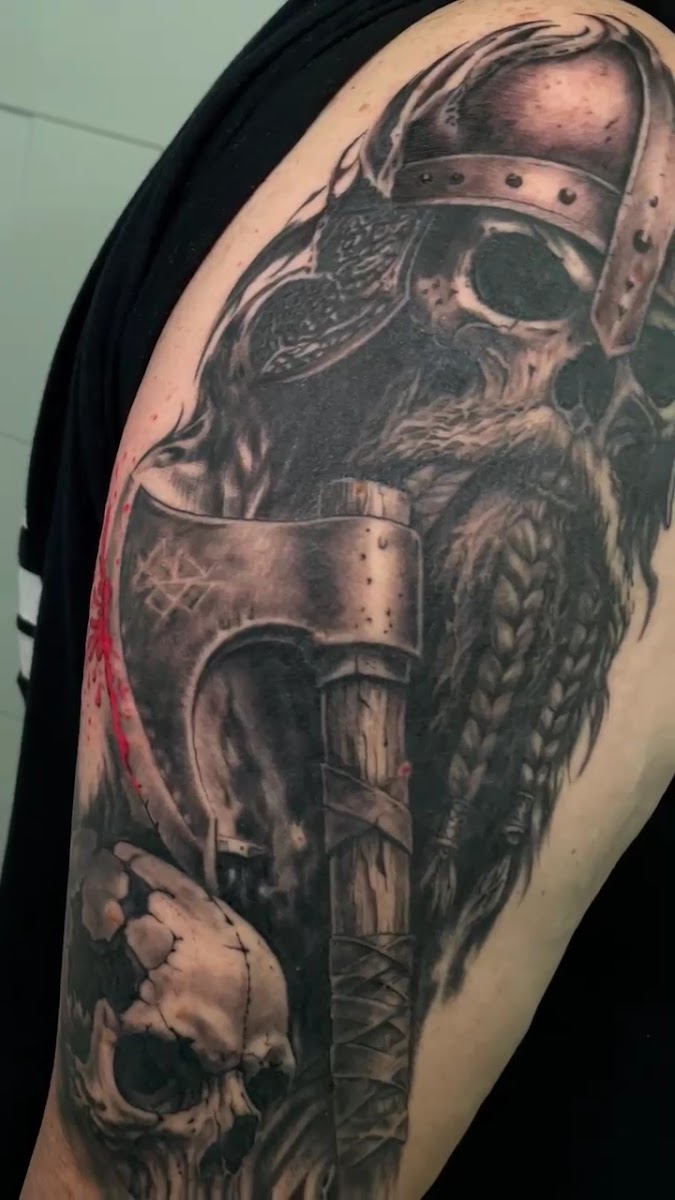 Brazo tatuado en negro y gris con un guerrero de casco remachado y barba trenzada, un hacha con runas en la hoja y una calavera debajo.