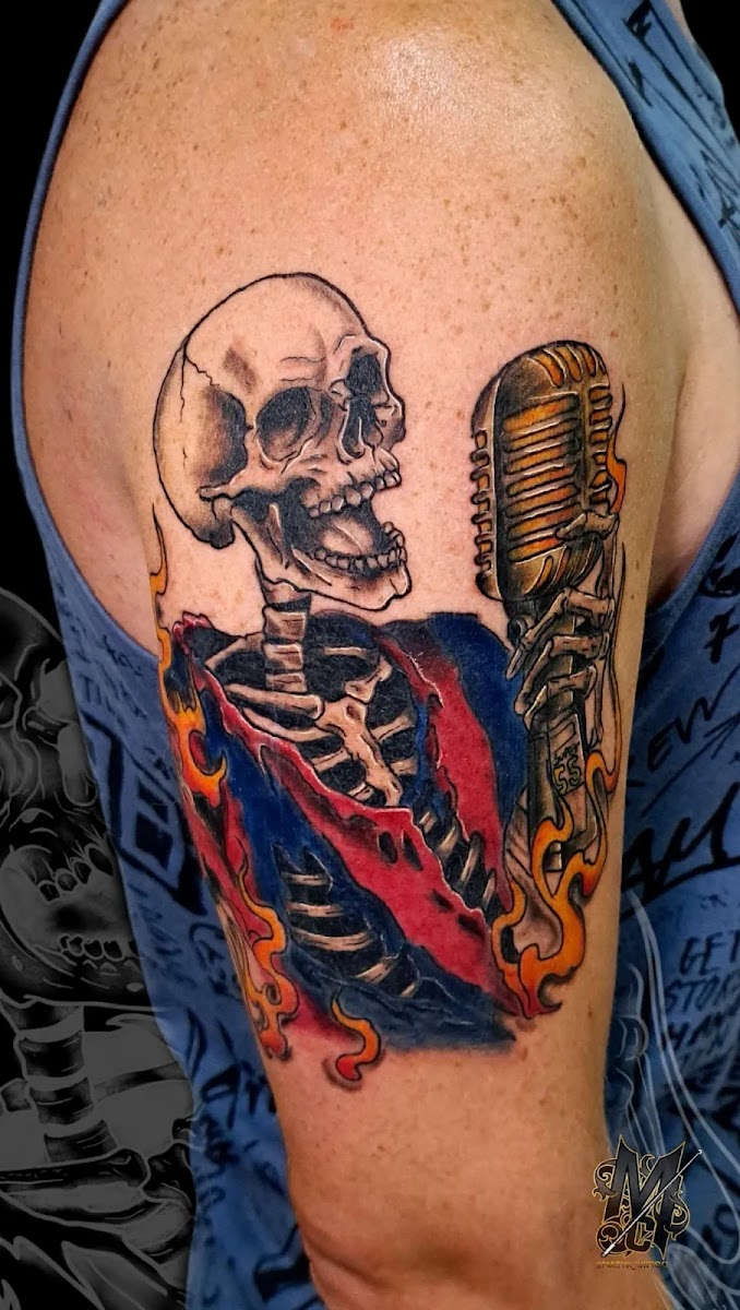 Brazo tatuado a color con un esqueleto cantando ante un micrófono de cinta dorado, envuelto en llamas naranjas y una tela roja y azul.
